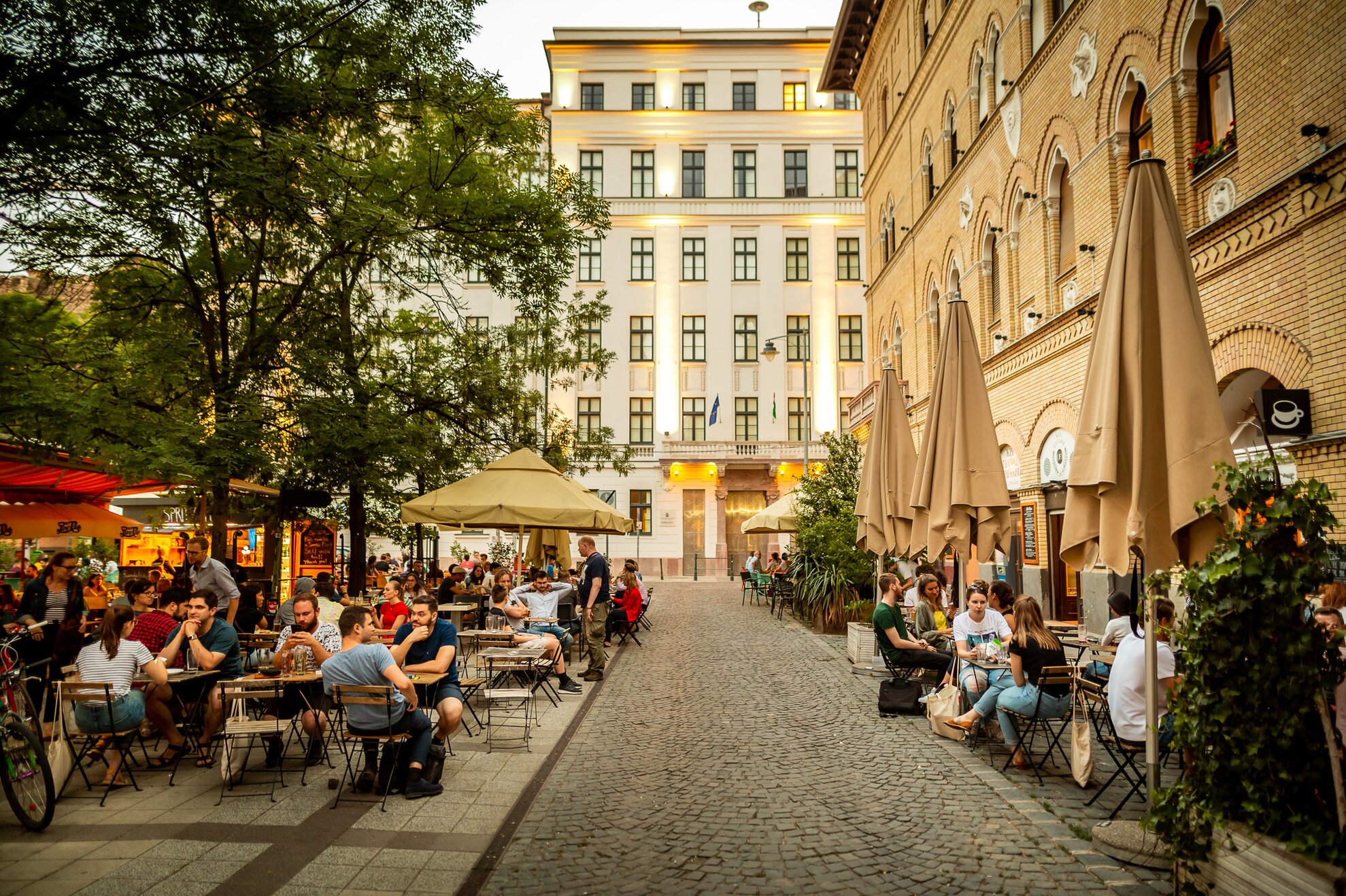 Cobblestone street, trees and bustling terraces in front of illuminated buildings in Józsefváros.